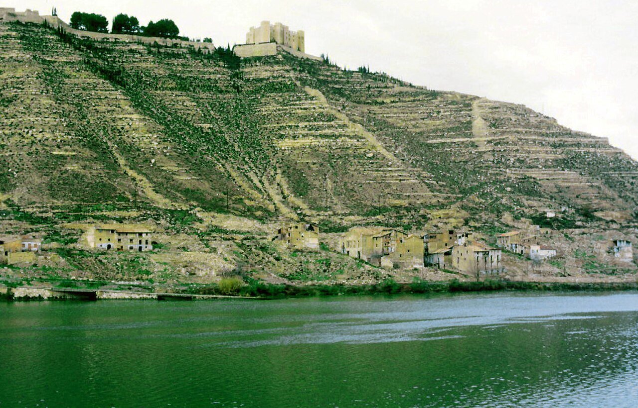 Casas abandonadas del pueblo viejo de Mequinenza en una ladera de bancales, con el castillo arriba y el agua verde del embalse abajo.