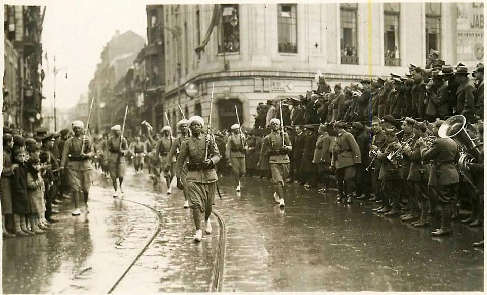 Soldados con turbante y fusil al hombro desfilan por una calle mojada de Gijón; a la derecha toca una banda militar y a la izquierda miran niños y vecinos desde la acera.